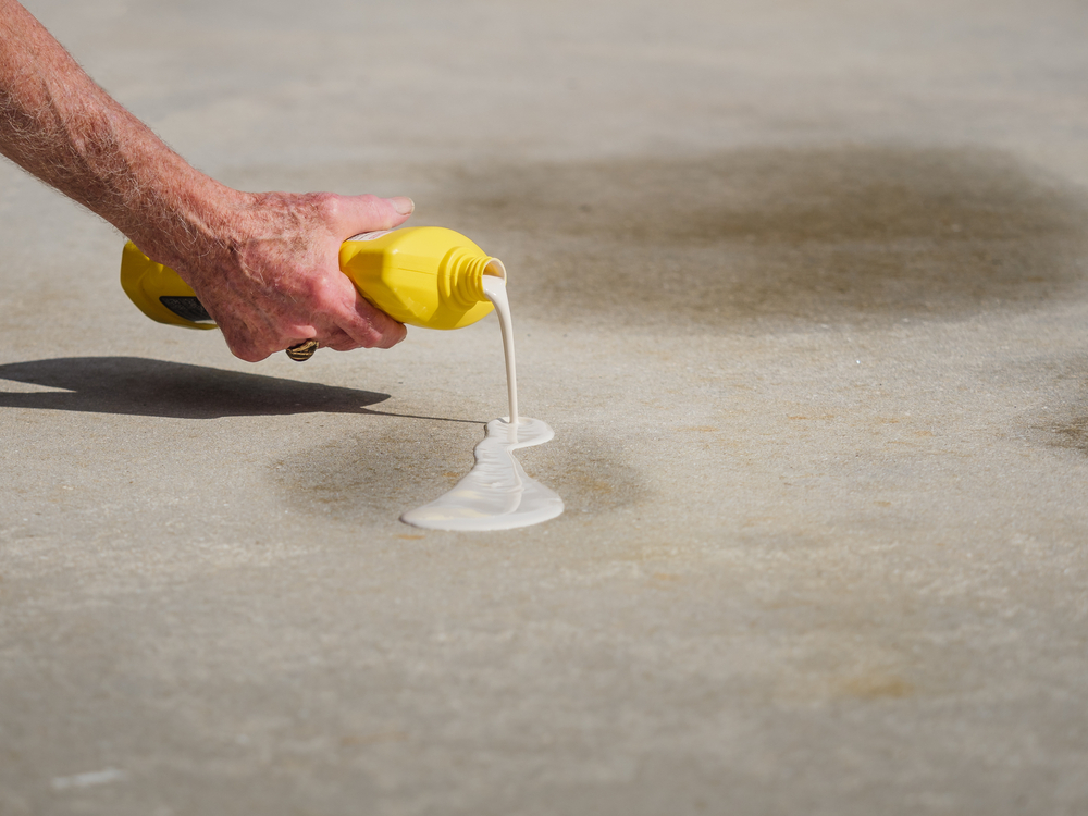 Hand pouring white liquid from a yellow bottle over an oil stain on a driveway